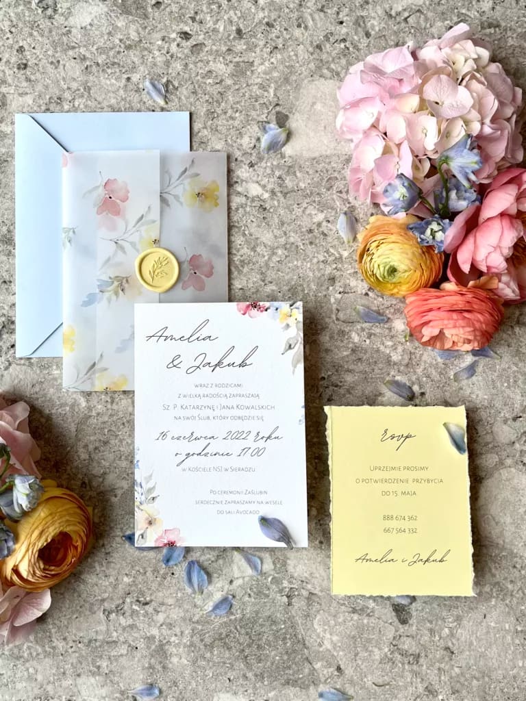Zaproszenie ślubne z kopertą kalkową oraz żółtą kartą RSVP, otoczone świeżymi kwiatami i płatkami w pastelowych kolorach.